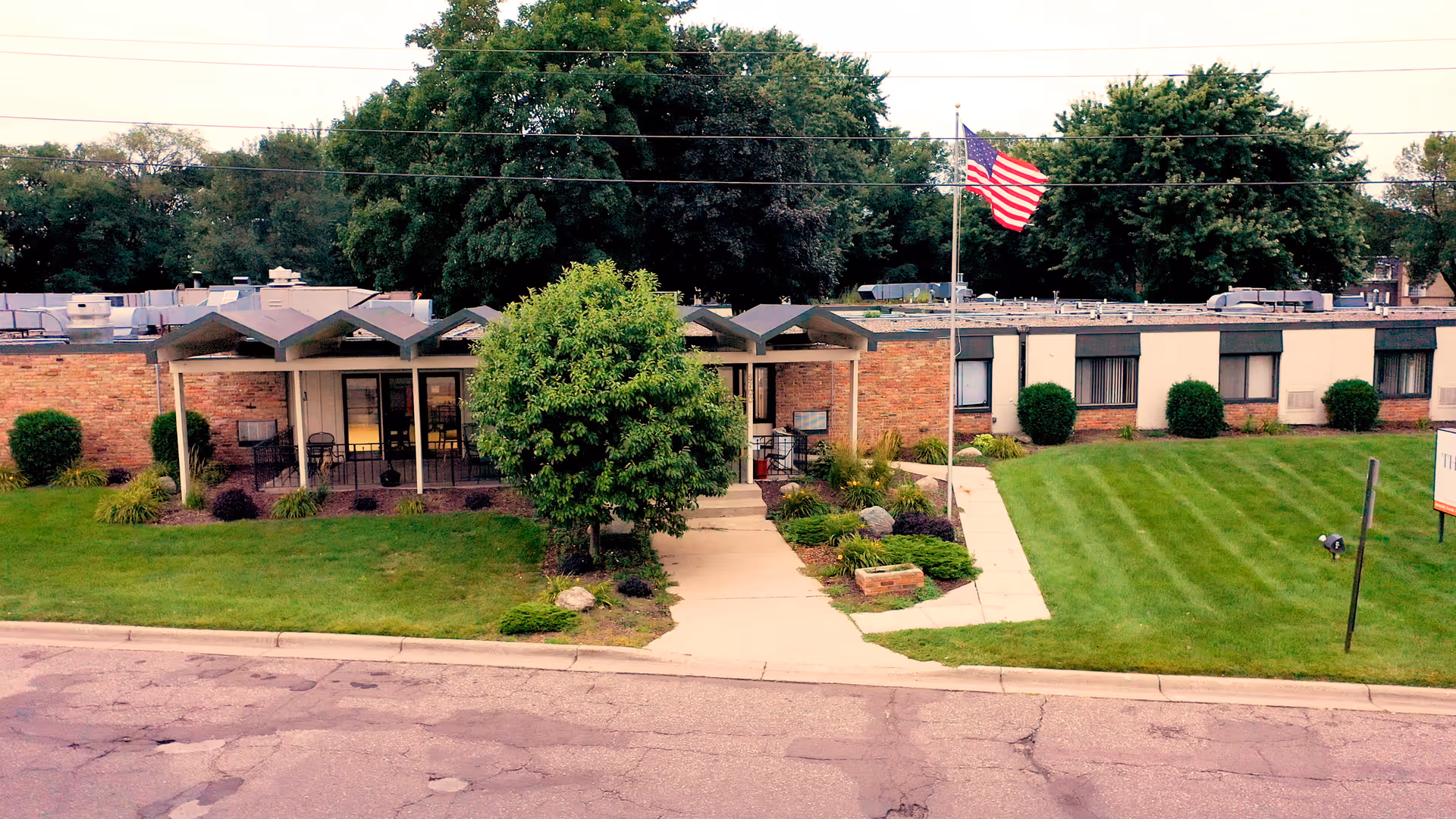 Exterior view of The Estates at Fridley facility showing a single-story brick building with a covered entrance, a flagpole with an American flag, well-maintained green lawns, bushes, and trees.