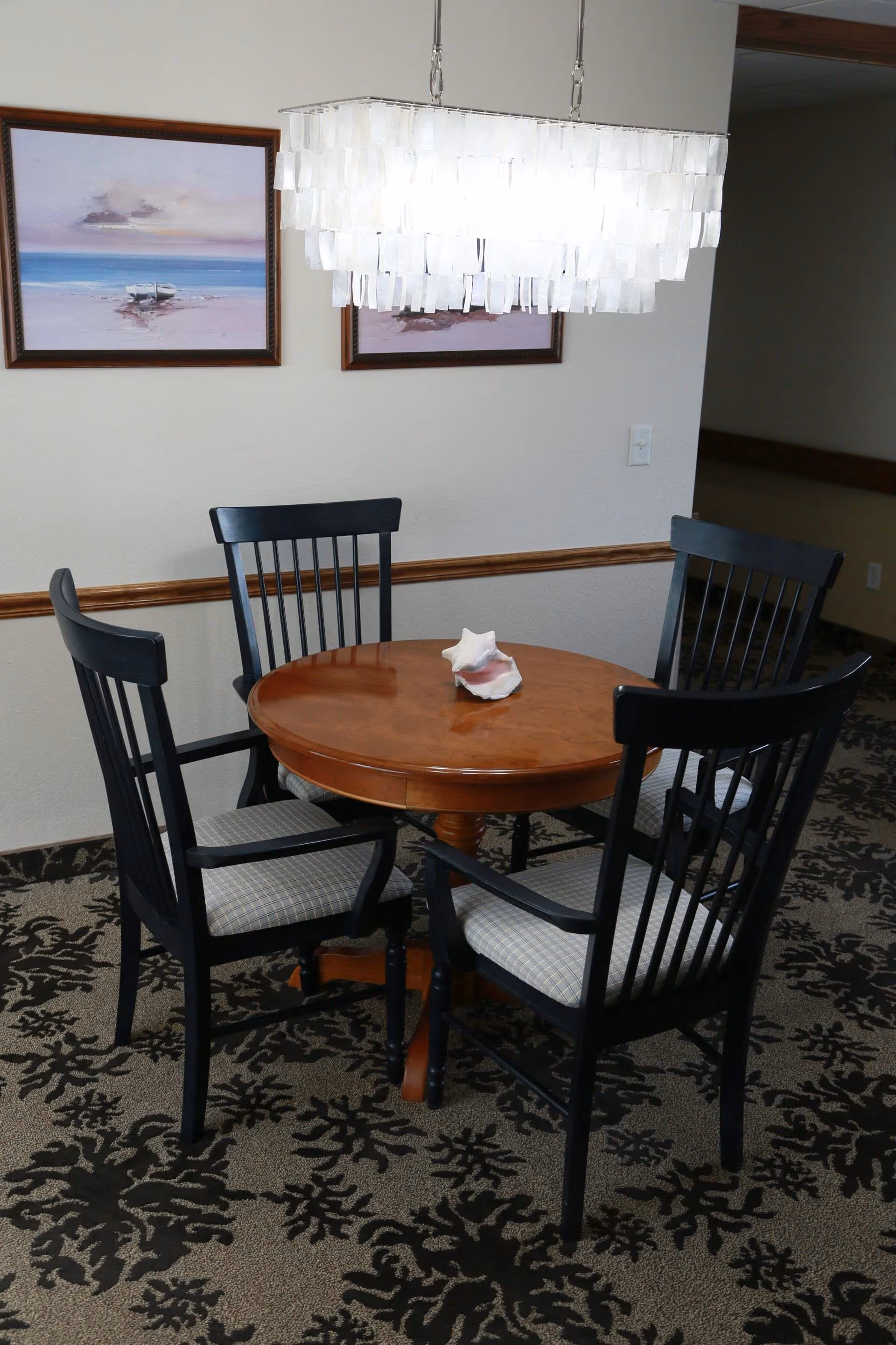 A small round wooden dining table with a decorative seashell centerpiece, surrounded by four black wooden chairs with cushioned seats. Above the table hangs a modern chandelier with rectangular translucent pieces. Two framed paintings of beach scenes are mounted on the wall behind the table.