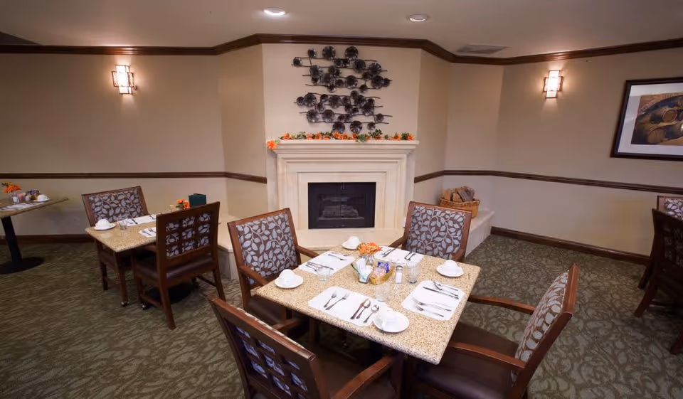 A cozy dining area with a square table set for four, featuring plates, cups, and silverware. The room has patterned carpet, beige walls with wooden trim, a fireplace decorated with a metal wall art piece above it, and soft wall sconces providing warm lighting. Additional tables and chairs are visible in the background along with a framed picture on the wall.