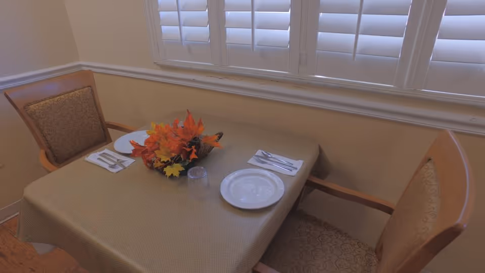 A small dining table set for two with a beige tablecloth, two chairs with patterned cushions, two plates, utensils on napkins, a clear drinking glass, and a centerpiece of autumn leaves in a cornucopia basket, positioned in front of a window with white plantation shutters.