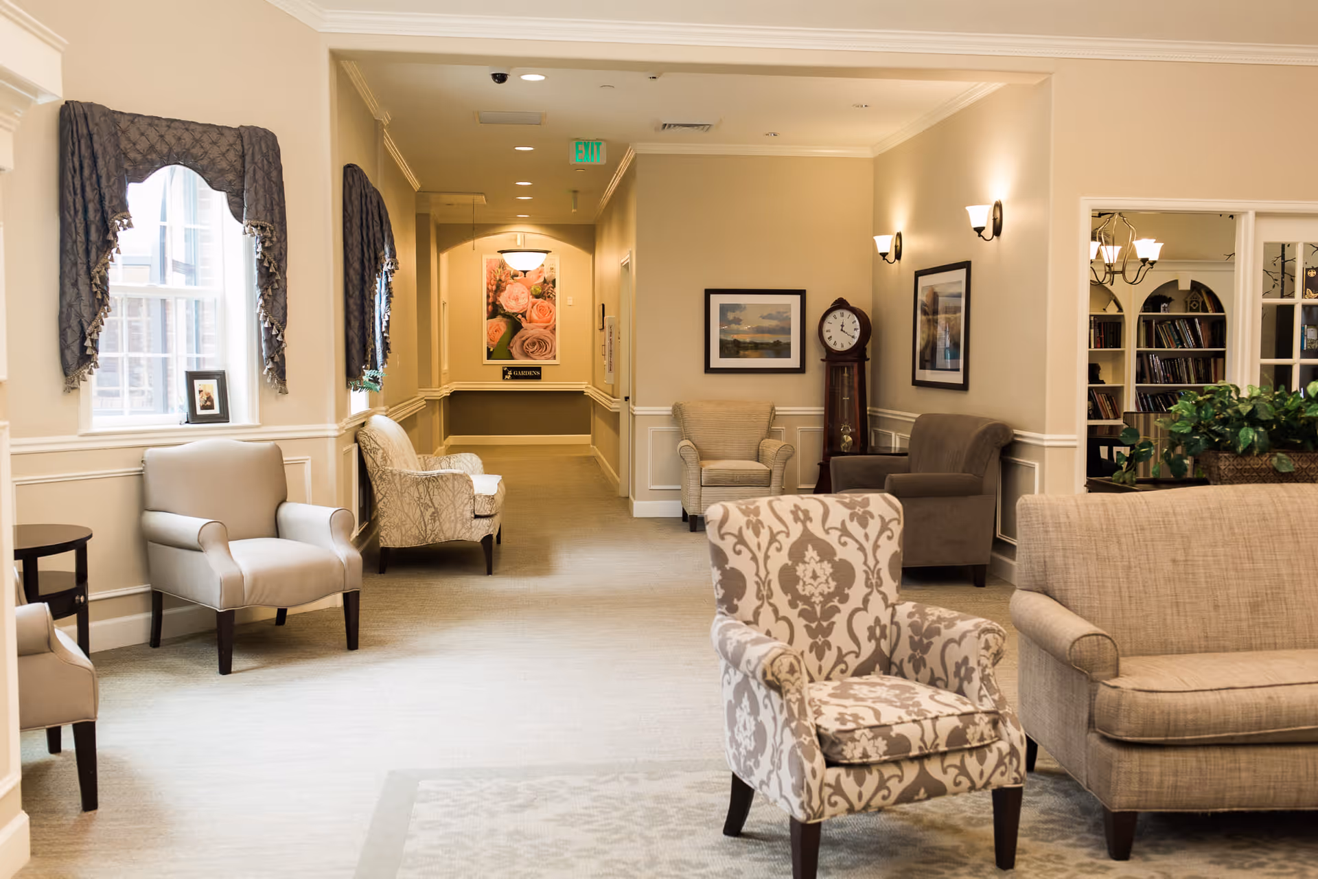 A well-lit, cozy sitting area in a senior living facility with several upholstered armchairs and a sofa arranged around a carpeted floor. The walls are painted beige with white trim, decorated with framed pictures and a grandfather clock. Windows with dark curtains allow natural light to enter, and a hallway with a floral picture is visible in the background.