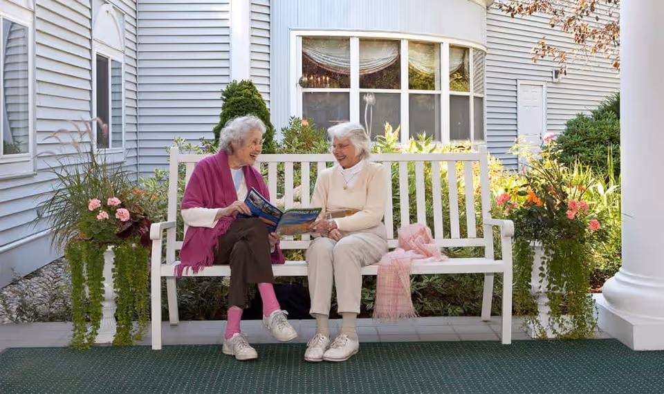 Two elderly women sitting on a white bench outside a building, engaged in conversation. One woman is wearing a purple shawl and holding a magazine, while the other is dressed in light-colored clothing. There are potted plants with flowers on either side of the bench, and the building has light gray siding with a large window behind them.