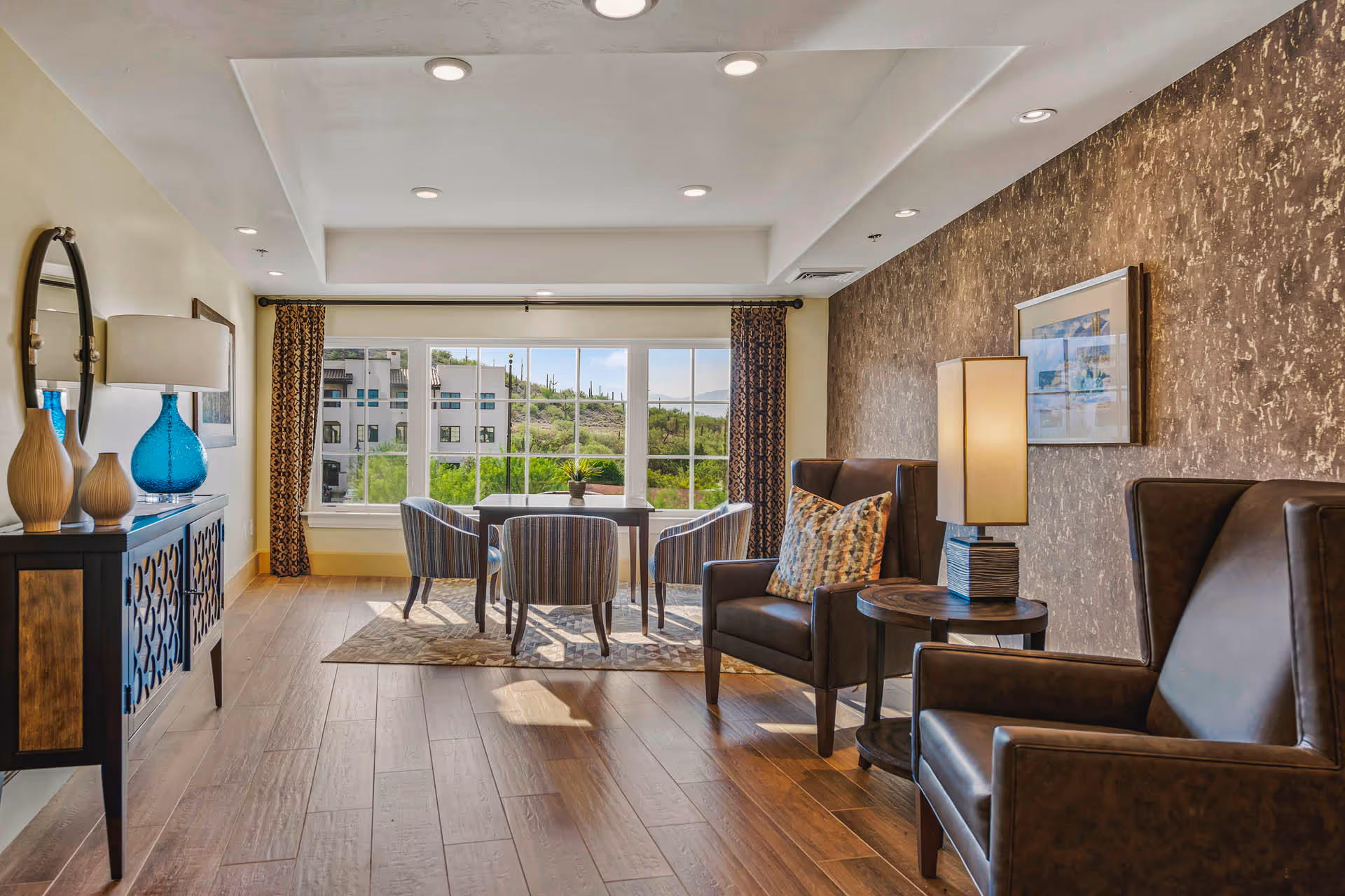 Sunlit communal lounge with wooden floors, armchairs and a small table and chairs by a large window overlooking greenery.