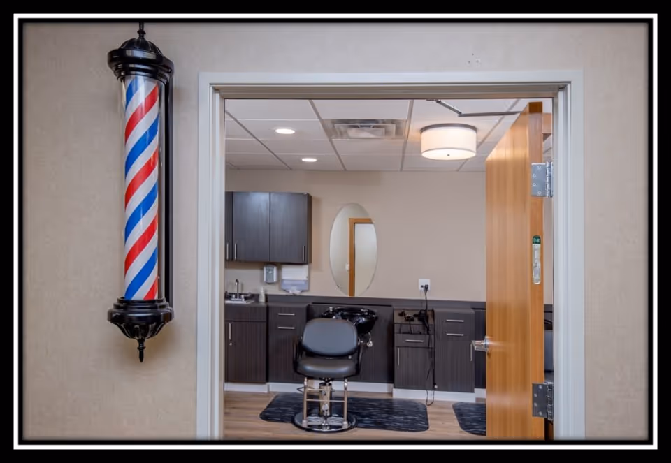 Interior view of a barber or salon room with a black barber chair in front of a counter with cabinets, a sink, and an oval mirror on the wall. A traditional red, white, and blue barber pole is mounted on the wall outside the room next to the door.