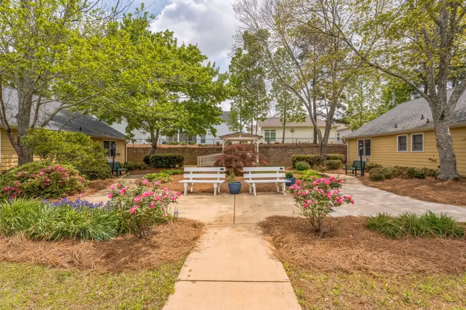 Outdoor courtyard area at Barclay House of Carrollton featuring a paved walkway leading to two white benches with a potted plant between them. The courtyard is surrounded by green trees, flowering bushes, and landscaped garden beds. Yellow buildings with windows and benches are visible on either side of the courtyard under a partly cloudy sky.