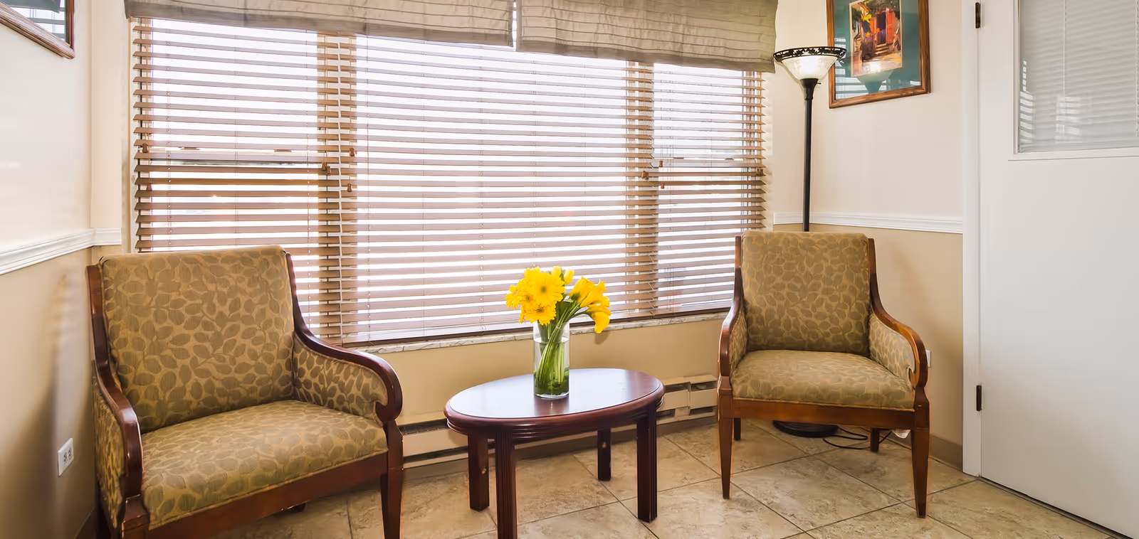 A small sitting area with two patterned armchairs facing each other and a round wooden table between them holding a vase with yellow flowers. Behind the chairs is a large window with wooden blinds partially closed. A floor lamp and a framed picture are visible on the right side near a white door.