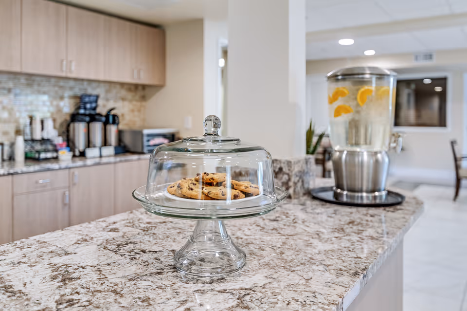A countertop with a glass cake stand holding chocolate chip cookies under a glass dome. In the background, there is a beverage dispenser with water and lemon slices, coffee dispensers, and a microwave in a kitchen or common area setting.