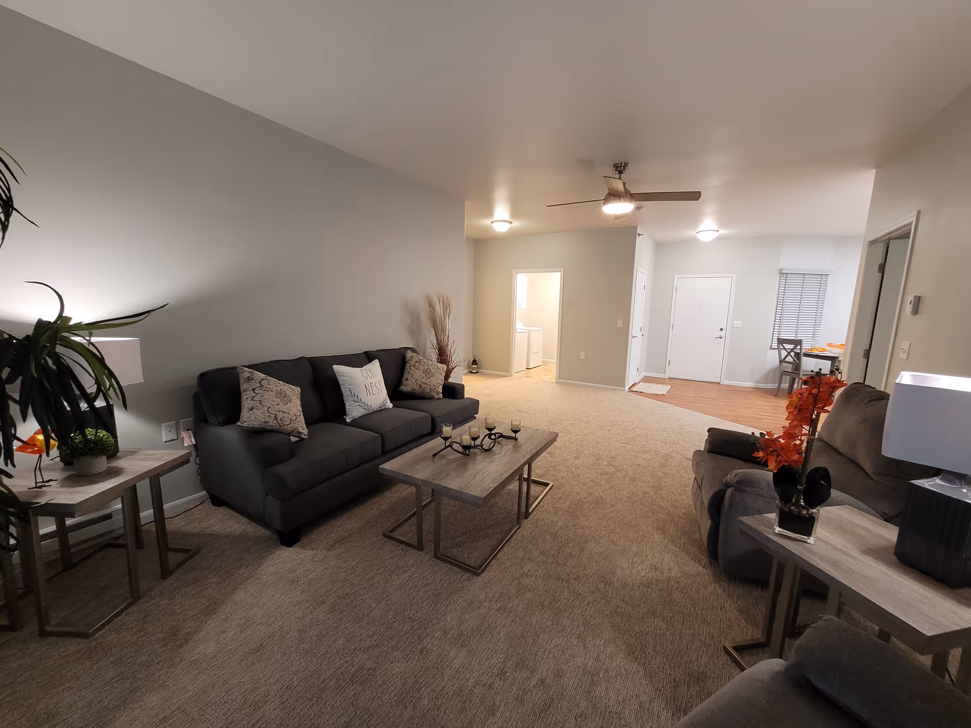 A spacious living room with beige carpet and light gray walls. The room features a dark gray sofa with decorative pillows, a matching armchair, and a wooden coffee table with candle holders. There are side tables with lamps and plants on either side of the seating area. In the background, a hallway leads to a laundry room with a washer and dryer, and a dining area with a table and chairs is visible near the entrance door.