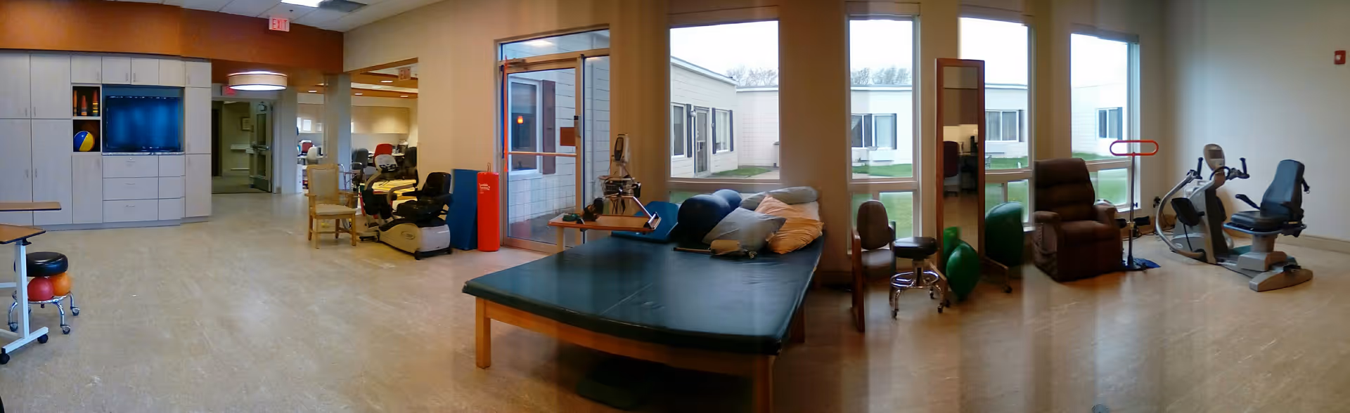 A spacious physical therapy or rehabilitation room with exercise equipment including stationary bikes, therapy balls, a padded therapy table with pillows, chairs, and large windows letting in natural light. The room has light-colored flooring and walls, with a view of an outdoor courtyard through the windows.