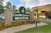 Entrance area of Camellia Gardens of Life Care facility with a large green and white sign displaying the facility name, surrounded by flowers and landscaping, and part of the building visible in the background.