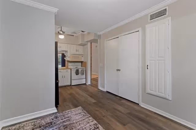 Interior view of a small kitchen area with white cabinets, a white stove, microwave, and refrigerator. The kitchen opens into a hallway with wooden flooring and light gray walls. There is a closed double-door closet on the right side of the hallway.