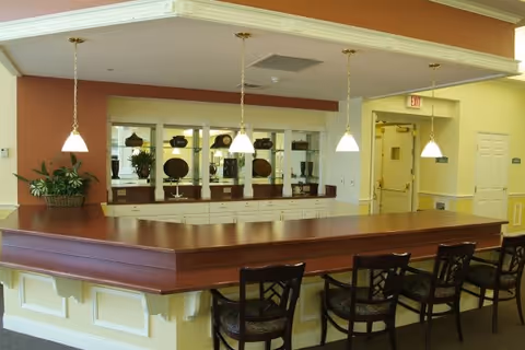 Interior view of a senior living facility dining or common area with a large wooden counter surrounded by four chairs. Above the counter, three pendant lights hang from the ceiling. Behind the counter, there is a mirrored wall with decorative plates and plants, and to the right, a hallway with doors and an exit sign is visible.