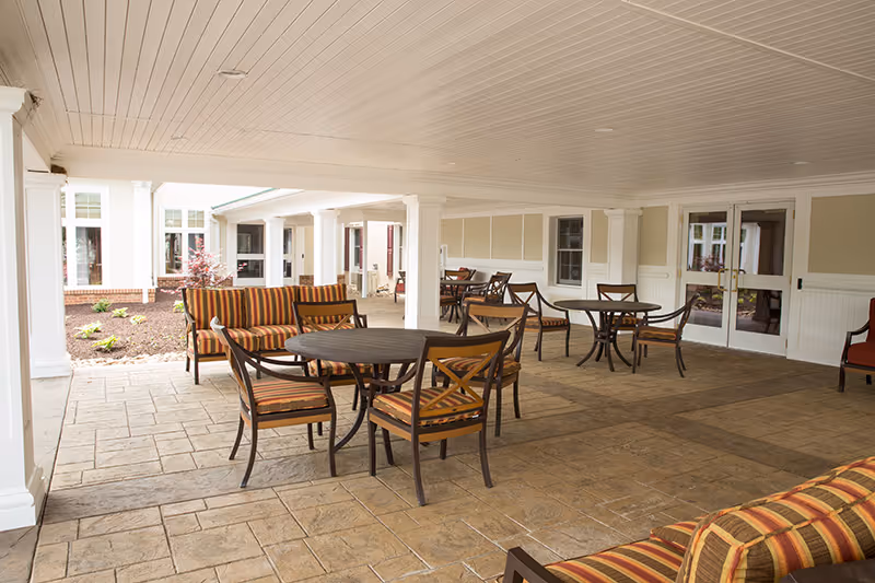 Covered outdoor seating area with multiple round tables and chairs featuring striped cushions, adjacent to a garden bed with small plants and a building with large windows and double doors.