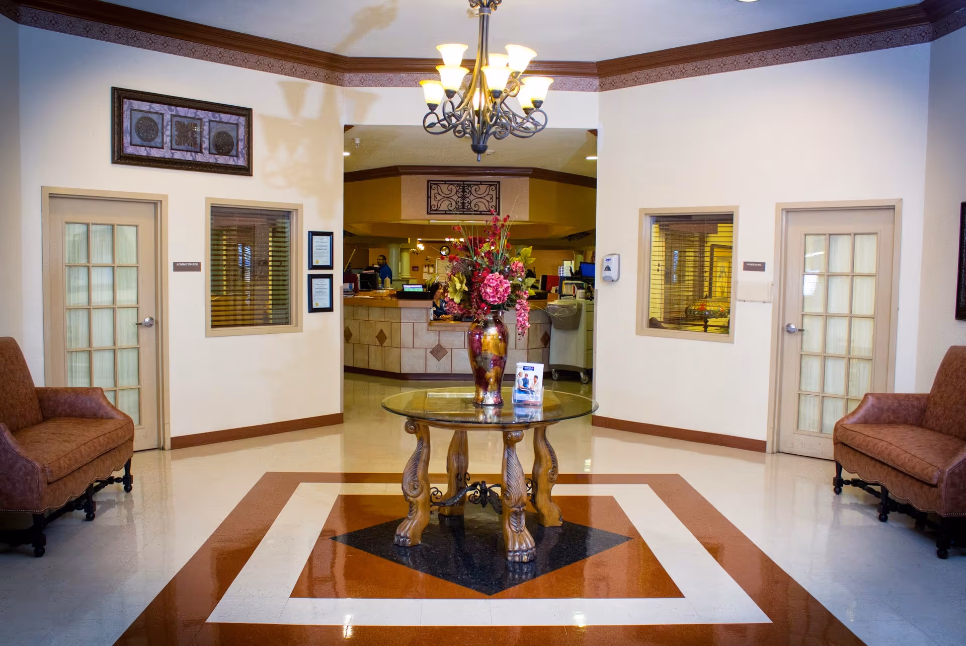 A well-lit interior lobby area with a decorative glass-top table holding a large floral arrangement in the center. Two upholstered chairs are positioned on either side against cream-colored walls with wooden trim. There are two doors with glass panels and windows on either side of the central opening leading to a reception or nurse station area in the background. A chandelier hangs from the ceiling above the table.