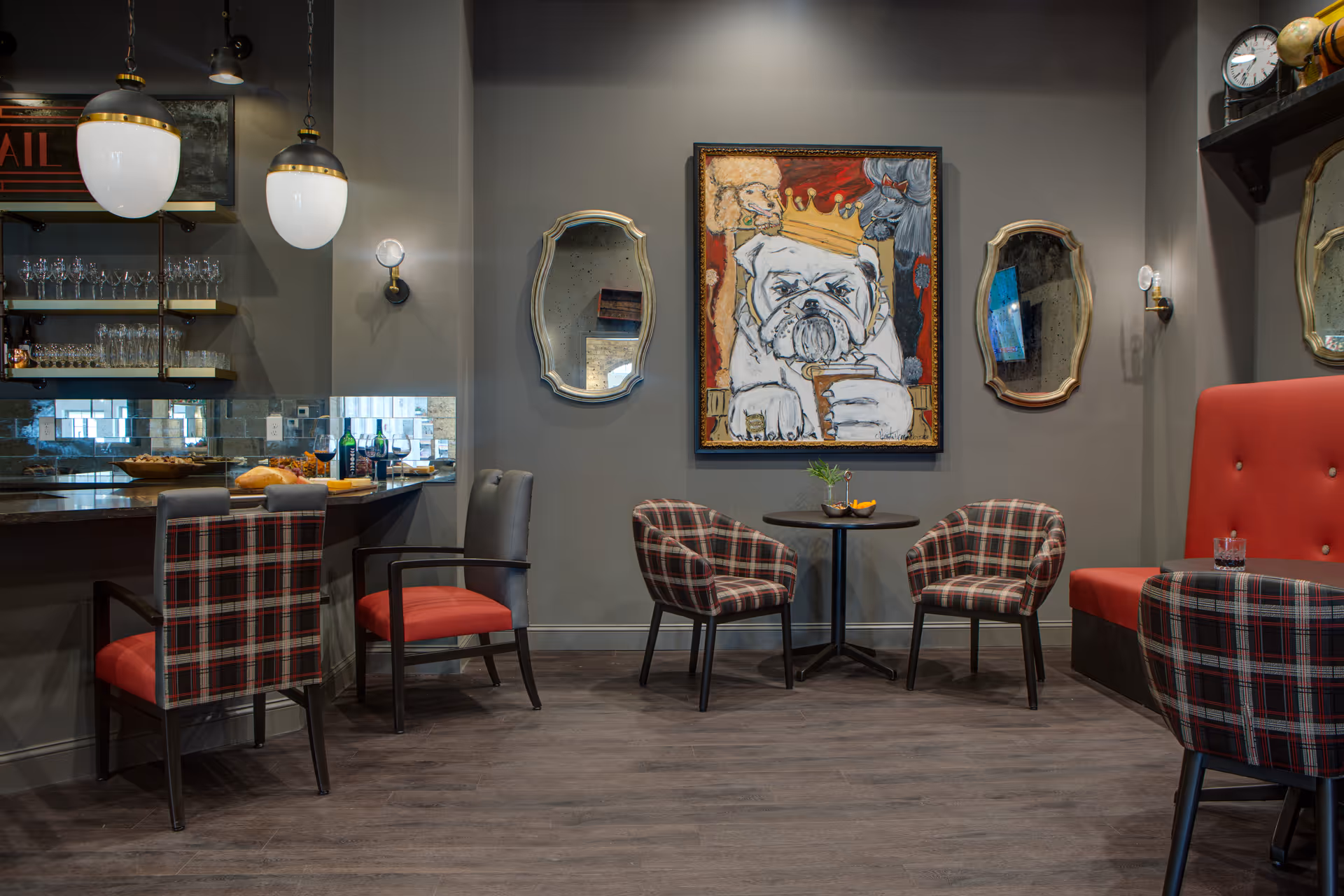 A cozy interior seating area with plaid upholstered chairs and red cushioned seats around small round tables. The walls are painted gray and decorated with two ornate mirrors and a colorful painting of a bulldog wearing a crown. There is a bar counter with shelves holding glasses and bottles, illuminated by hanging pendant lights.