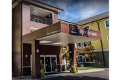 Entrance of Marquis Piedmont Post Acute Rehab facility showing a covered drop-off area with the facility name and 'Assisted Living' signage. The building exterior is visible with windows and a yellow section of the building in the background under a partly cloudy sky.