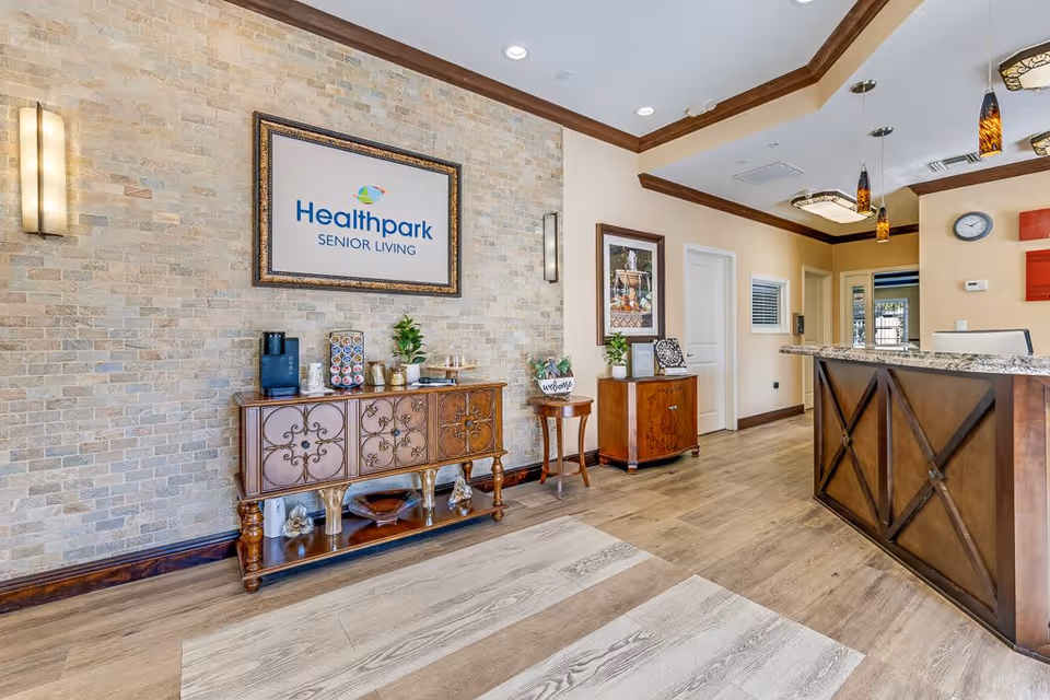 Reception area of Healthpark Senior Living featuring a wooden front desk with granite countertop, decorative pendant lights, a framed Healthpark Senior Living sign on a brick accent wall, and wooden furniture with plants and coffee supplies.