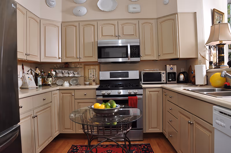 A well-lit kitchen with beige cabinets, stainless steel appliances including a microwave and stove, a glass-top round table with a bowl of lemons and limes, and a window with a lamp on the countertop beside a double sink.