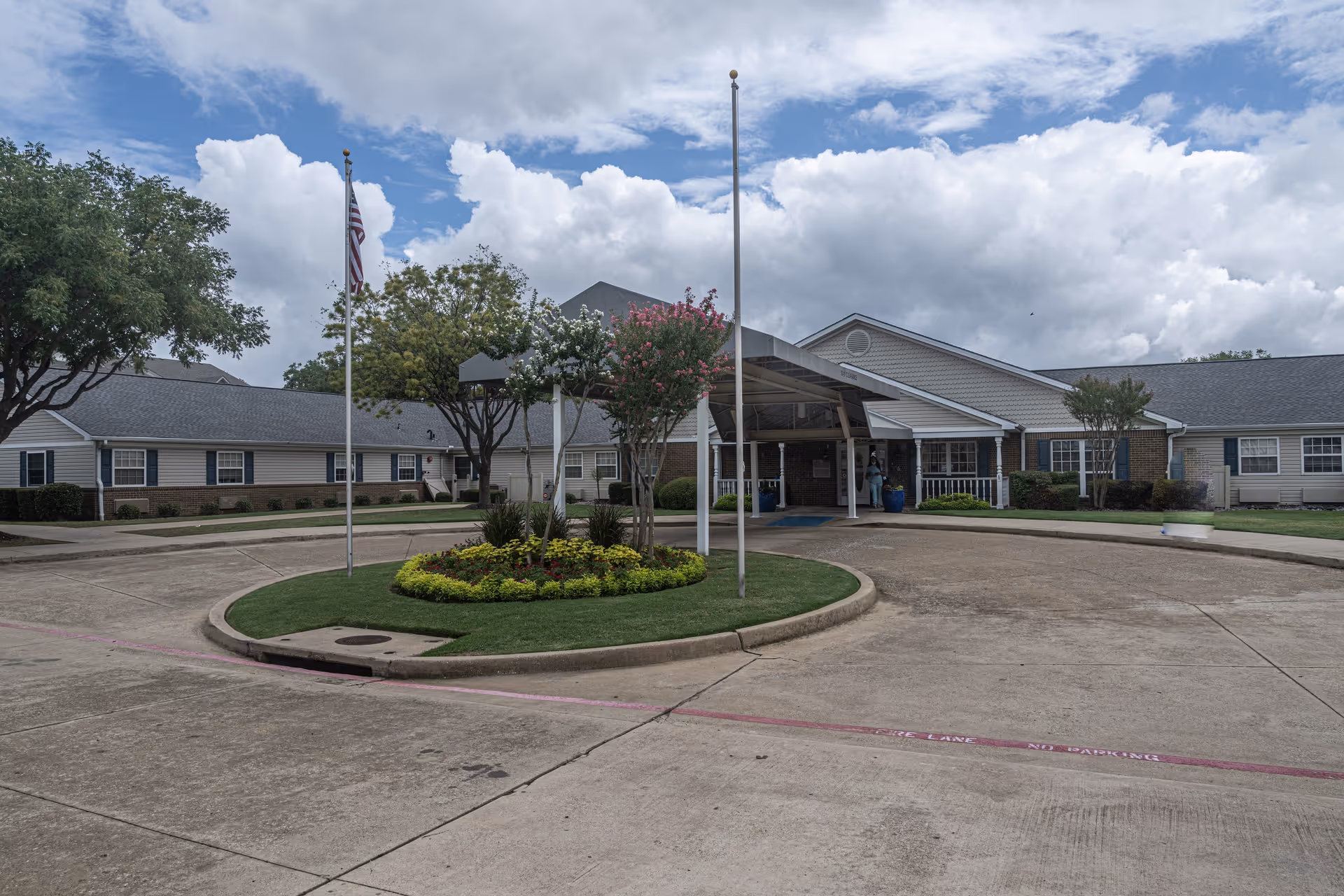 Exterior view of Arden Courts A ProMedica Memory Care Community in Richardson showing a circular driveway with a landscaped island in the center, two flagpoles, and the entrance to the building under a covered drop-off area. The building is single-story with a combination of brick and siding, surrounded by trees and shrubbery under a partly cloudy sky.