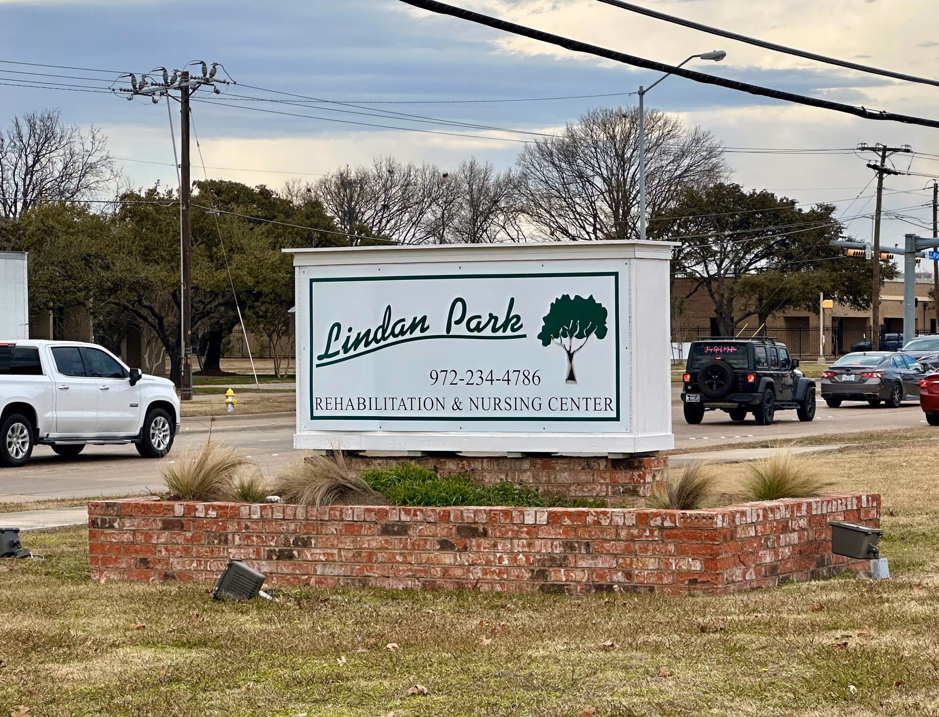 A roadside sign for Lindan Park Rehabilitation & Nursing Center, mounted on a brick base with some grass and plants around it. Several cars are visible on the road behind the sign, along with trees and utility poles under a cloudy sky.