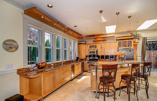 Bright kitchen with wooden cabinets, granite countertops, and a large island with three high chairs. The kitchen features stainless steel appliances including a refrigerator, oven, and microwave. Multiple windows along one wall let in natural light, and there are pendant lights hanging above the island. A bowl of fruit and a vase with flowers are on the countertops.