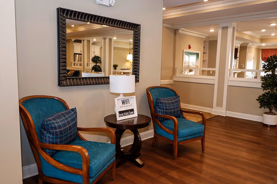 A cozy seating area with two teal upholstered wooden armchairs, each with a plaid cushion, positioned on either side of a small round wooden table holding a lamp and a sign that reads 'WIFI password home sweet home'. A large decorative mirror hangs on the beige wall behind the chairs, reflecting part of the room. The floor is wooden, and there is a potted plant in the corner.