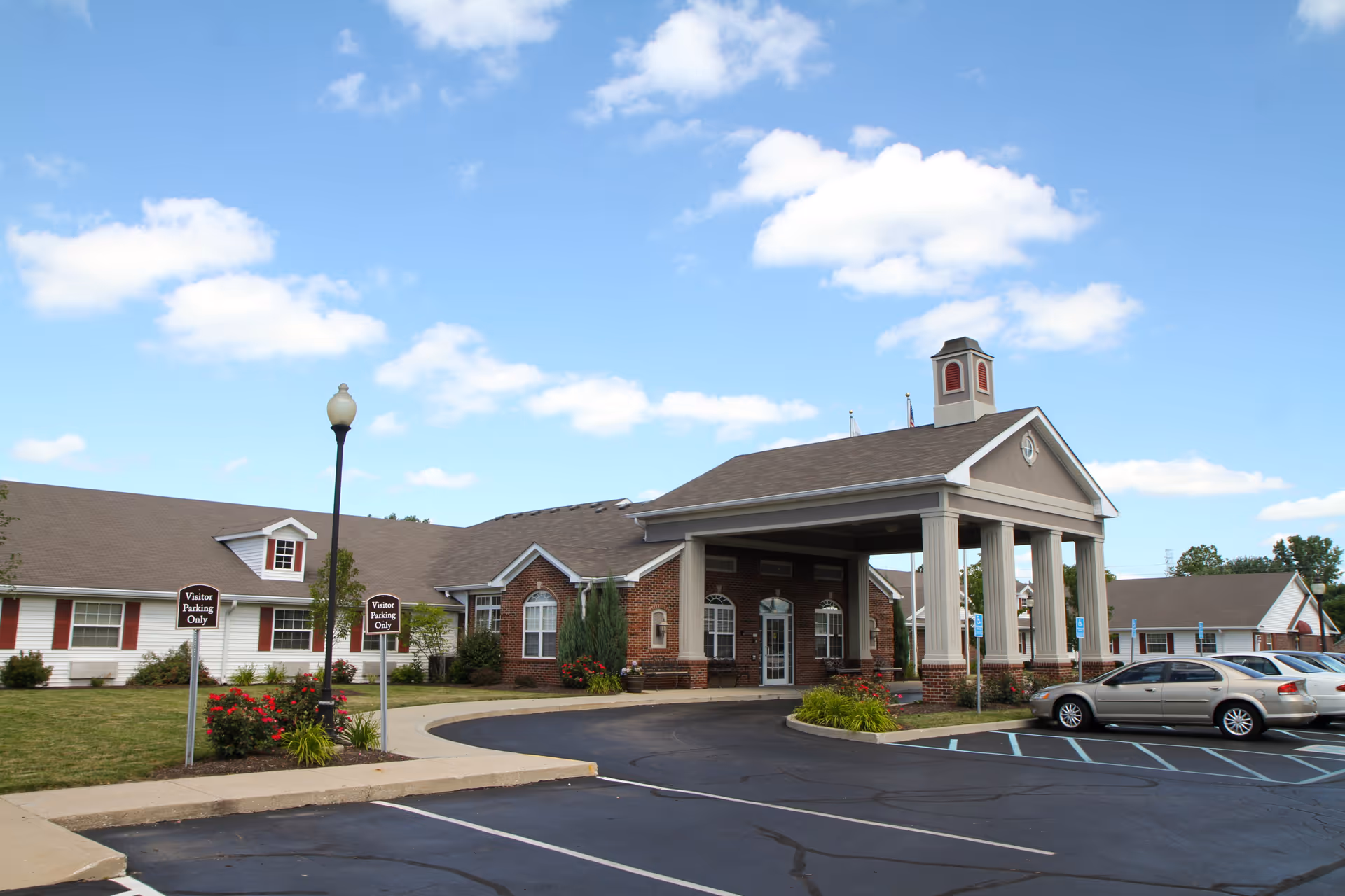 Exterior view of Five Star Residences of Banta Pointe showing a large building with a covered entrance supported by columns, a parking lot with several cars, and clear blue sky with some clouds.