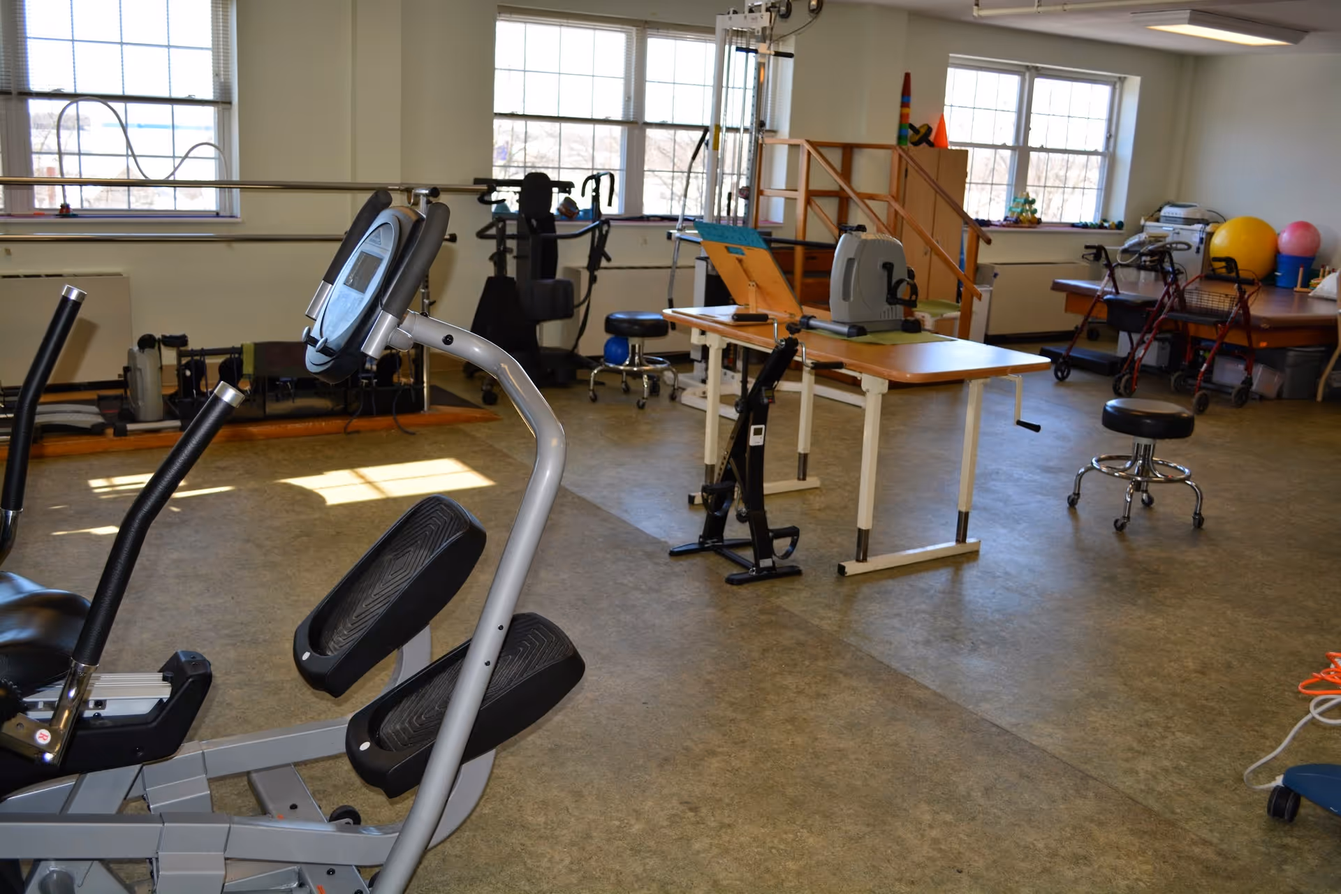 A bright rehabilitation gym room with exercise machines, therapy tables, walkers, and therapy balls by windows.