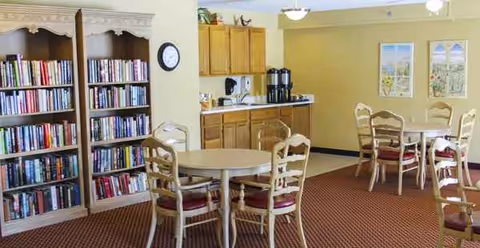 Interior common area with round tables, chairs, a bookshelf, plus kitchenette in a senior living facility.