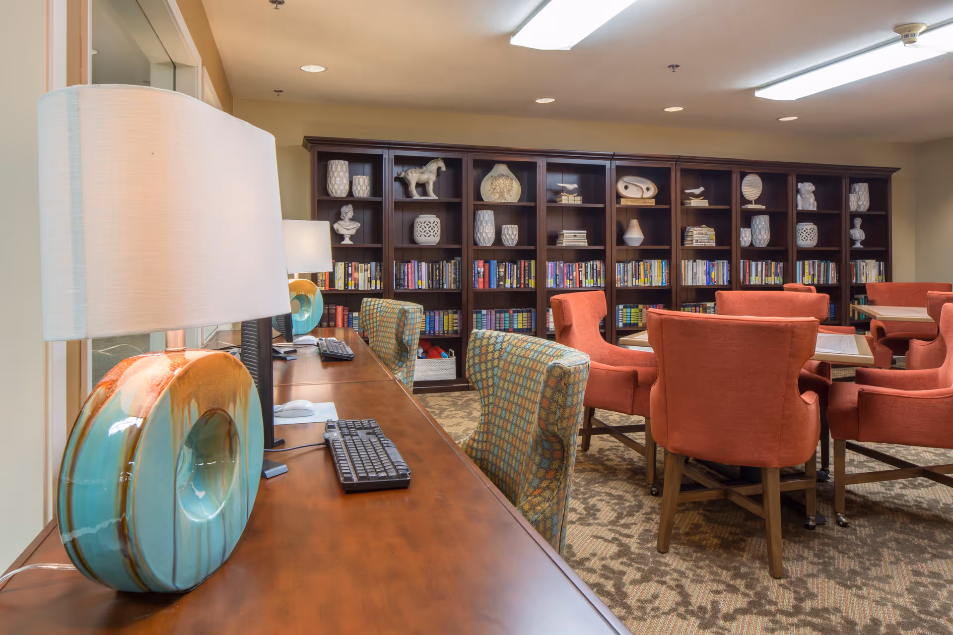 A well-lit room with a long wooden desk featuring computer monitors, keyboards, and decorative lamps with turquoise bases. In the background, there are bookshelves filled with books and decorative items. Several orange upholstered chairs surround tables, and patterned chairs are placed at the desk.