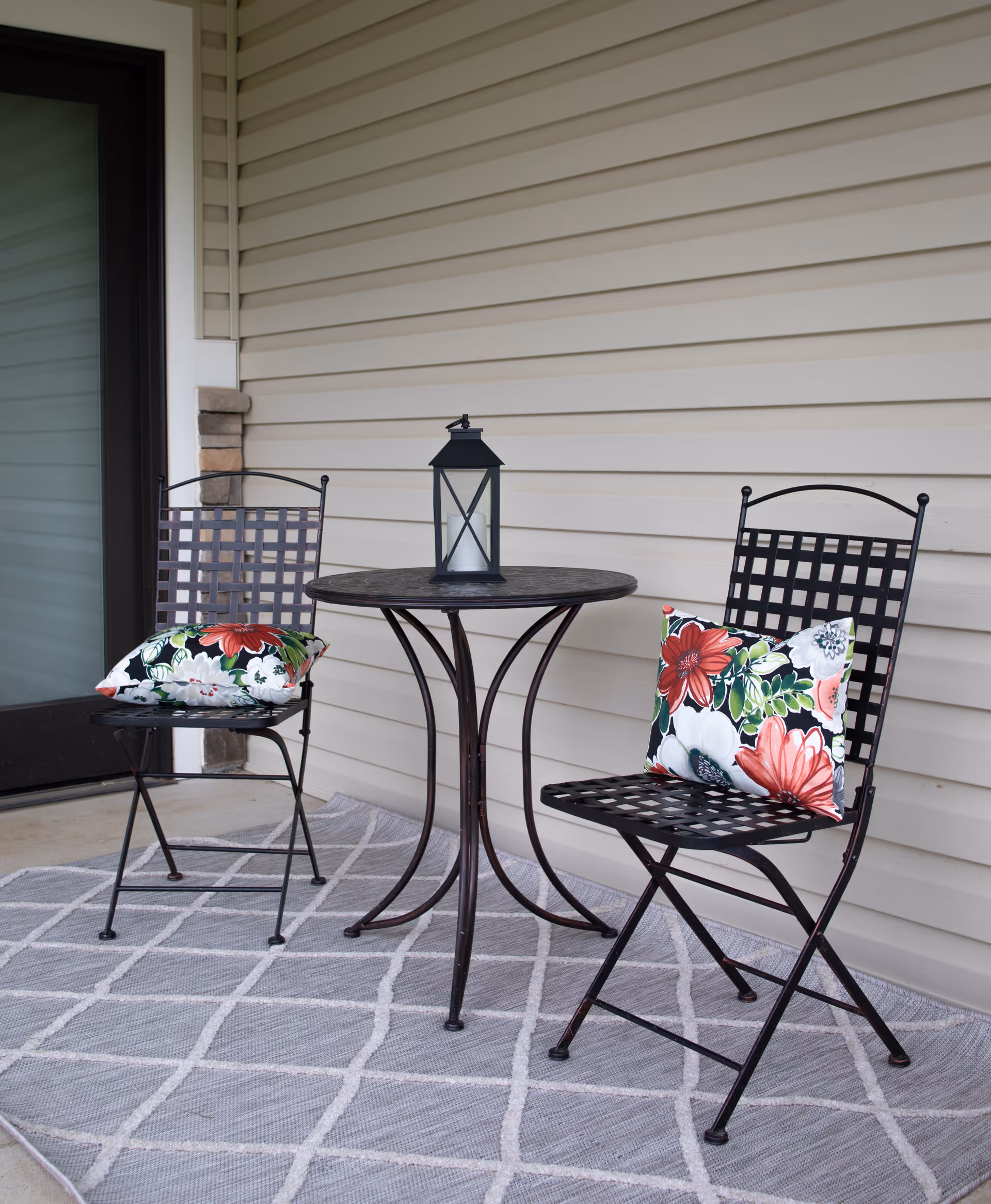 Small outdoor patio area with two black metal chairs featuring lattice backs and seats, each with a colorful floral cushion. Between the chairs is a round black metal table with a decorative lantern on top. The patio floor is covered with a gray and white patterned rug, and the background shows beige siding of the building.