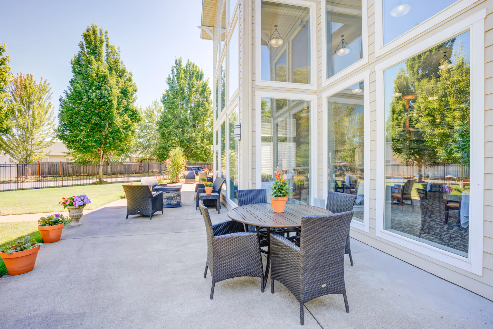 Outdoor patio area at Evergreen Senior Living Eugene with a round table and four wicker chairs, potted plants on the table and ground, large windows reflecting trees, and additional seating with a fire pit in the background under a clear sky.