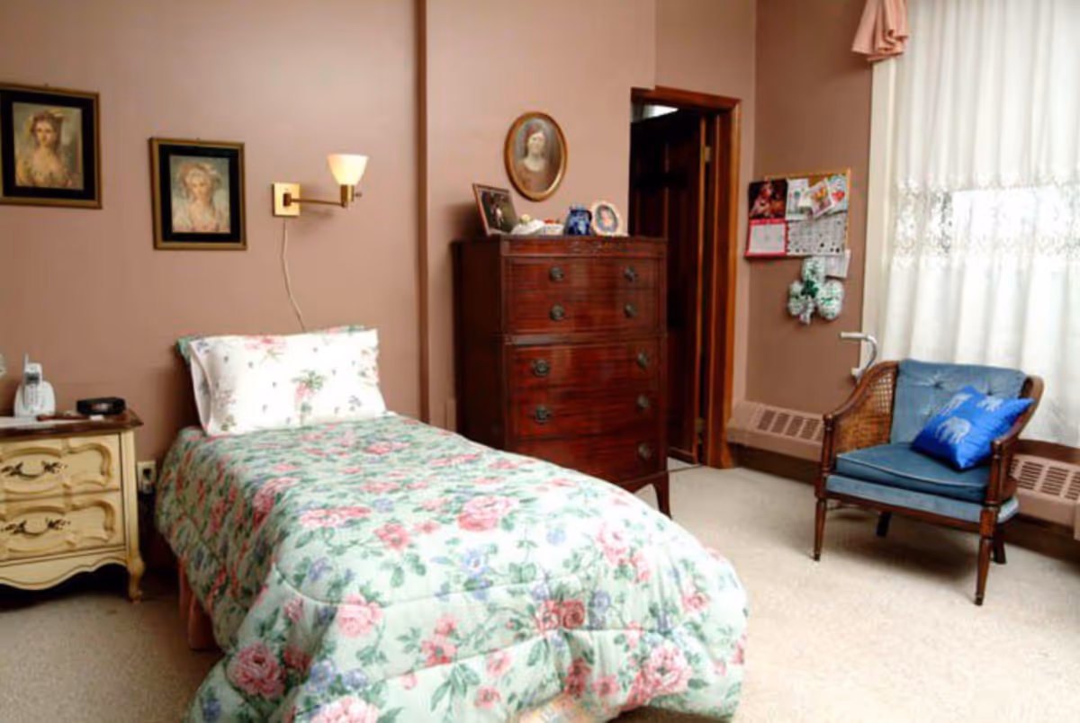 A cozy bedroom with a single bed covered in a floral quilt and matching pillow. The room features a wooden dresser with framed photos on top, a blue cushioned chair with a blue pillow, a nightstand with a phone and an ashtray, and framed portraits on the wall. A window with white lace curtains allows natural light into the room.
