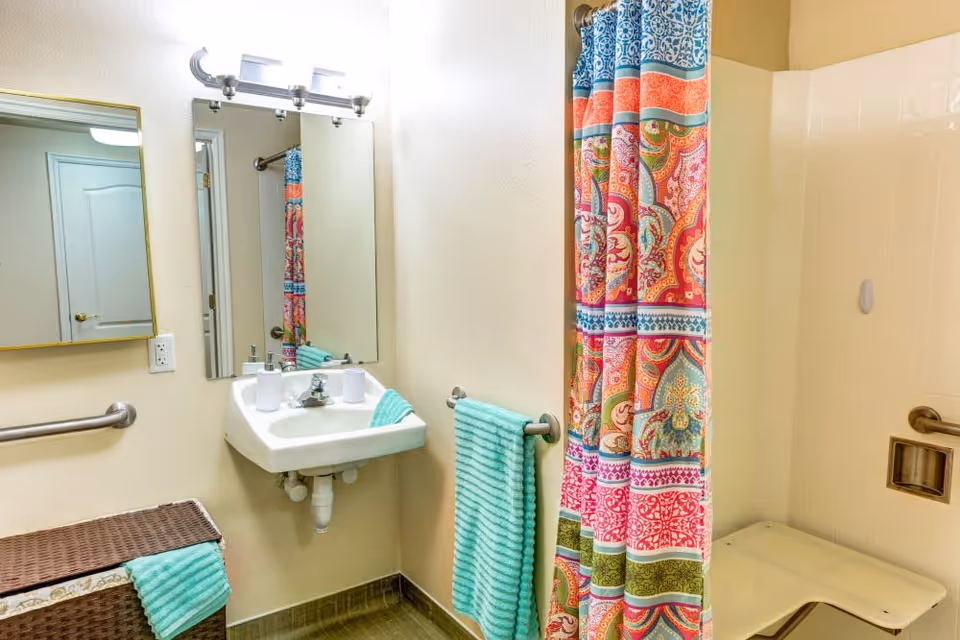 A bathroom with a white wall-mounted sink, a large mirror above it, and a light fixture with three bulbs. There is a colorful patterned shower curtain next to a beige tiled shower area with a built-in bench. A turquoise towel hangs on a metal towel rack, and a wicker laundry basket with a matching turquoise towel is placed near the sink. The walls are light-colored, and there is a metal grab bar on the wall near the sink.