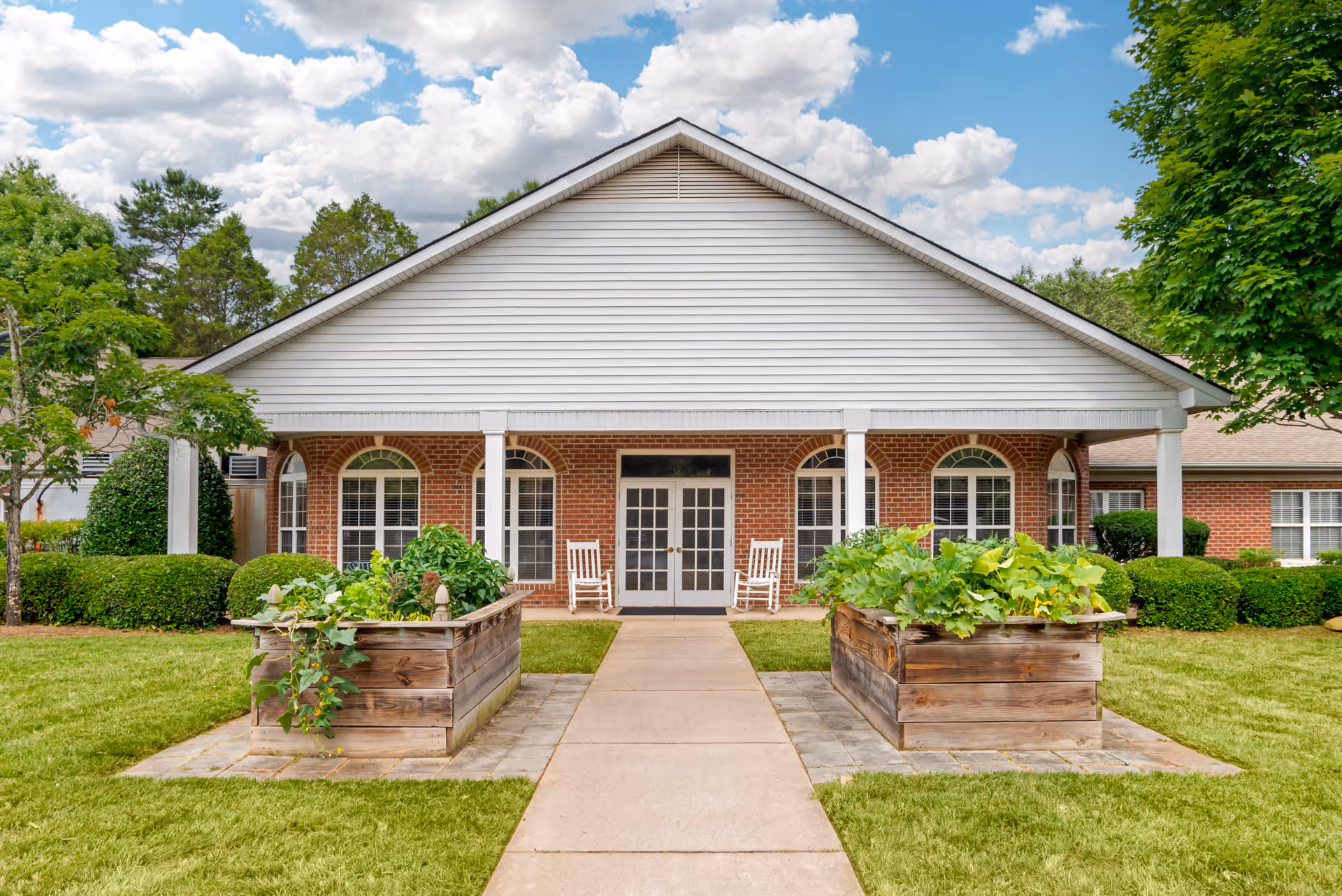 Front exterior view of a single-story brick building with white siding and a covered porch. The porch has two white rocking chairs and is flanked by two raised wooden garden beds filled with green plants. The building is surrounded by neatly trimmed bushes, trees, and a well-maintained lawn under a partly cloudy sky.