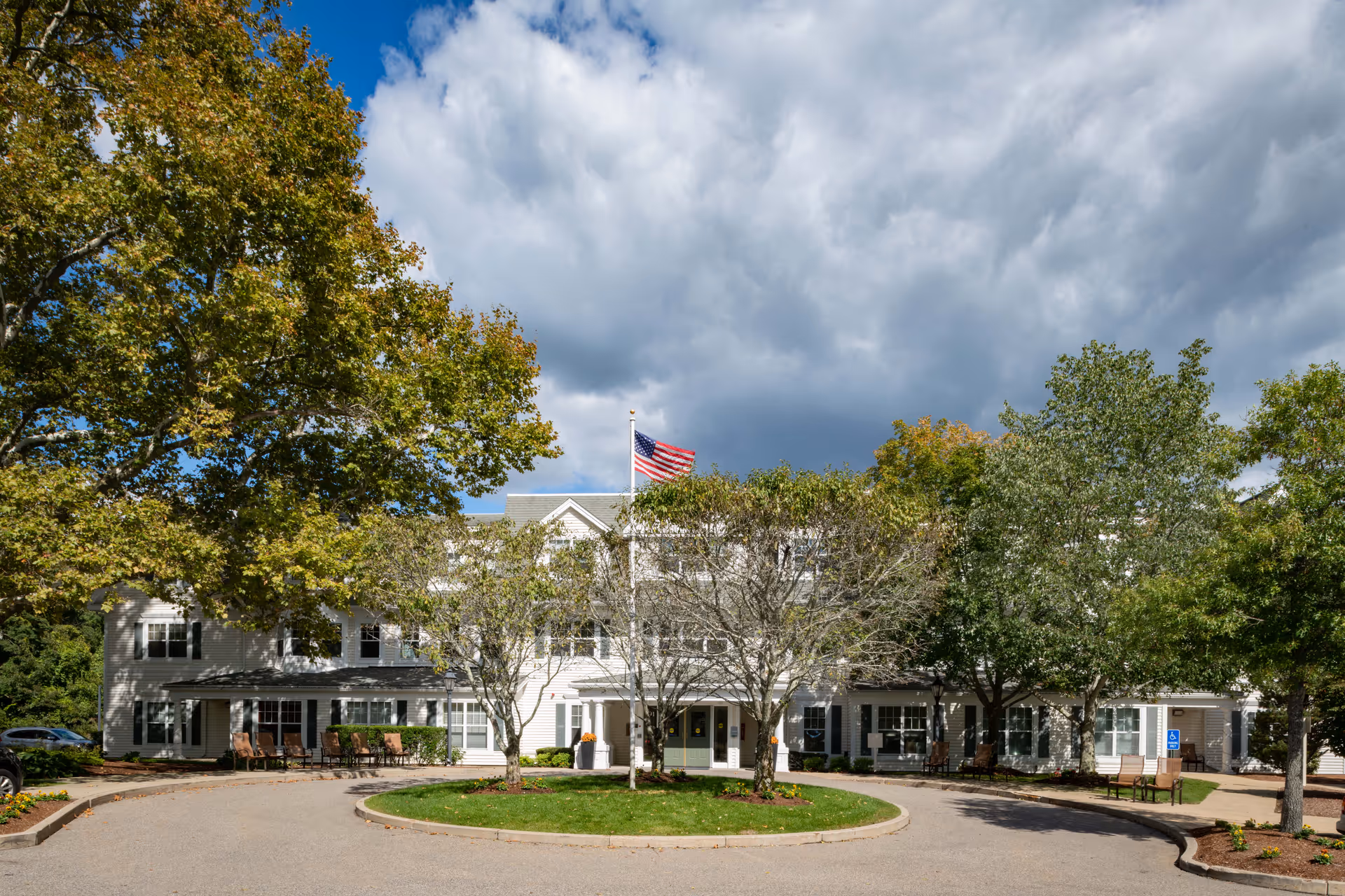 Front exterior of a two-story senior living facility with a circular driveway, trees, and an American flag flying above.