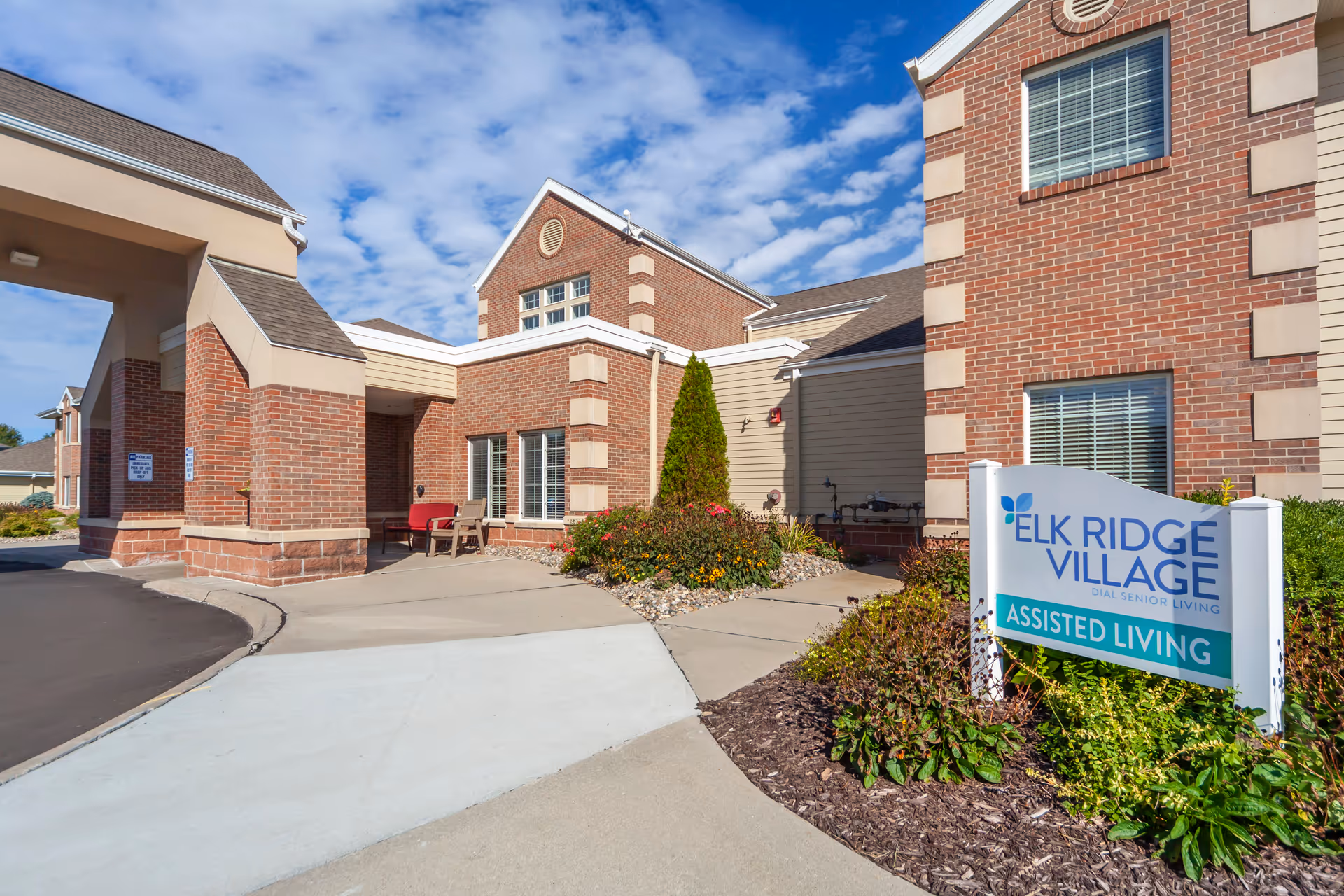 Exterior view of Elk Ridge Village Assisted Living facility showing a brick building with windows, a covered entrance with seating, landscaped bushes and flowers, and a sign reading 'Elk Ridge Village Assisted Living' under a partly cloudy blue sky.