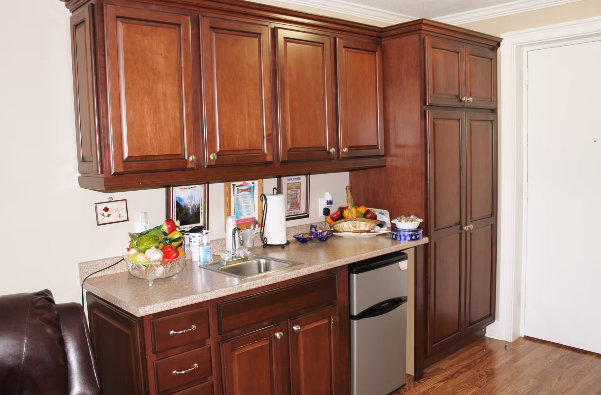 A kitchenette area with dark wooden cabinets, a small stainless steel sink, a mini refrigerator, and a countertop holding a basket of fruit, a bowl of snacks, a paper towel holder, and some other small items. The floor is wooden, and a white door is visible on the right side.