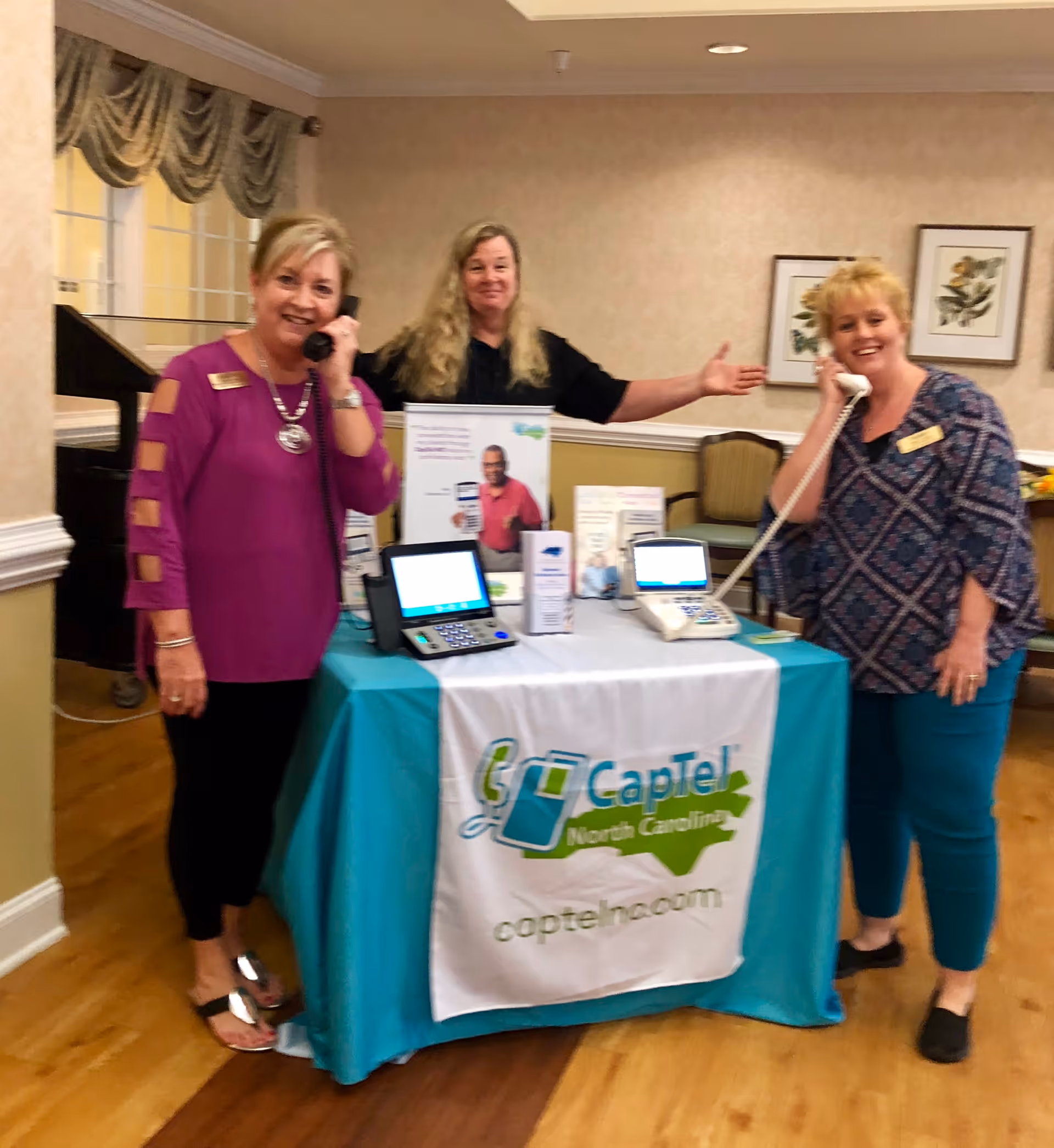 Three women standing behind a table with a teal tablecloth and a white banner that reads 'CapTel North Carolina captelnc.com'. Two women on the sides are holding telephones to their ears and smiling, while the woman in the middle is gesturing towards the table. The table displays telecommunication devices and informational brochures. The setting appears to be an indoor common area with framed pictures on the wall and wooden flooring.