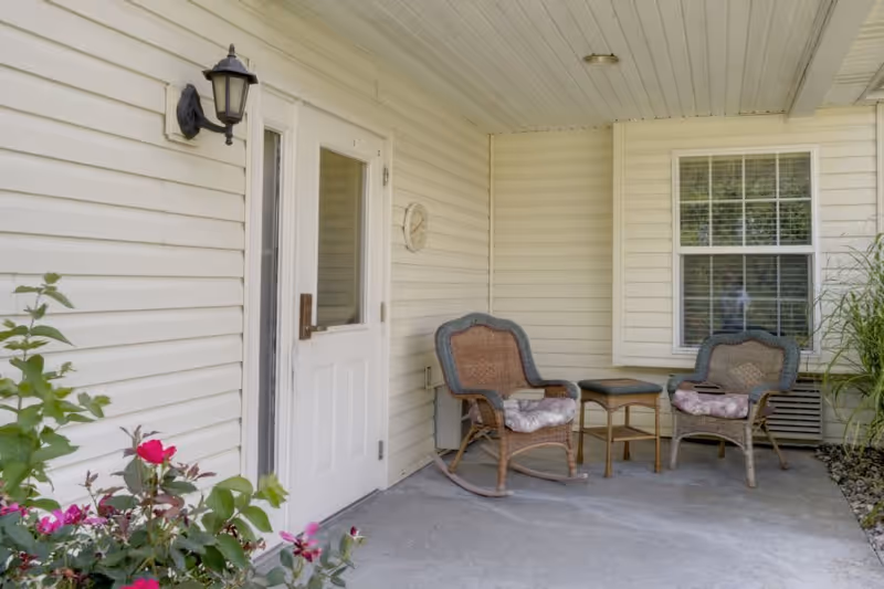 Covered outdoor patio area with two cushioned wicker rocking chairs and a small wicker table between them. There is a white door with a window, a wall-mounted lantern light, a clock on the wall, a window with white shutters, and some flowering plants in the foreground.