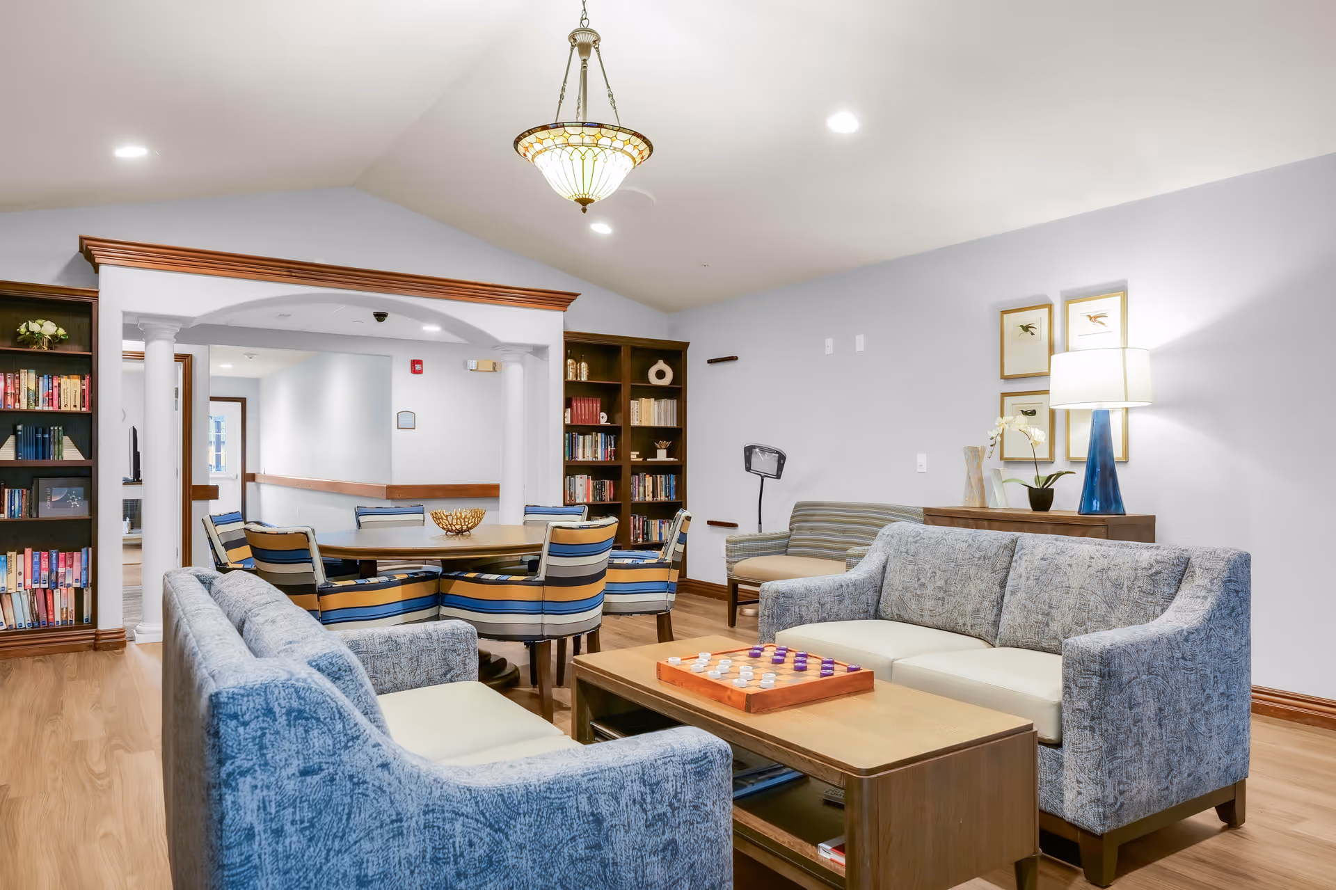 A bright communal living room with sofas, a coffee table holding a board game, bookshelves, and a dining table with striped chairs.