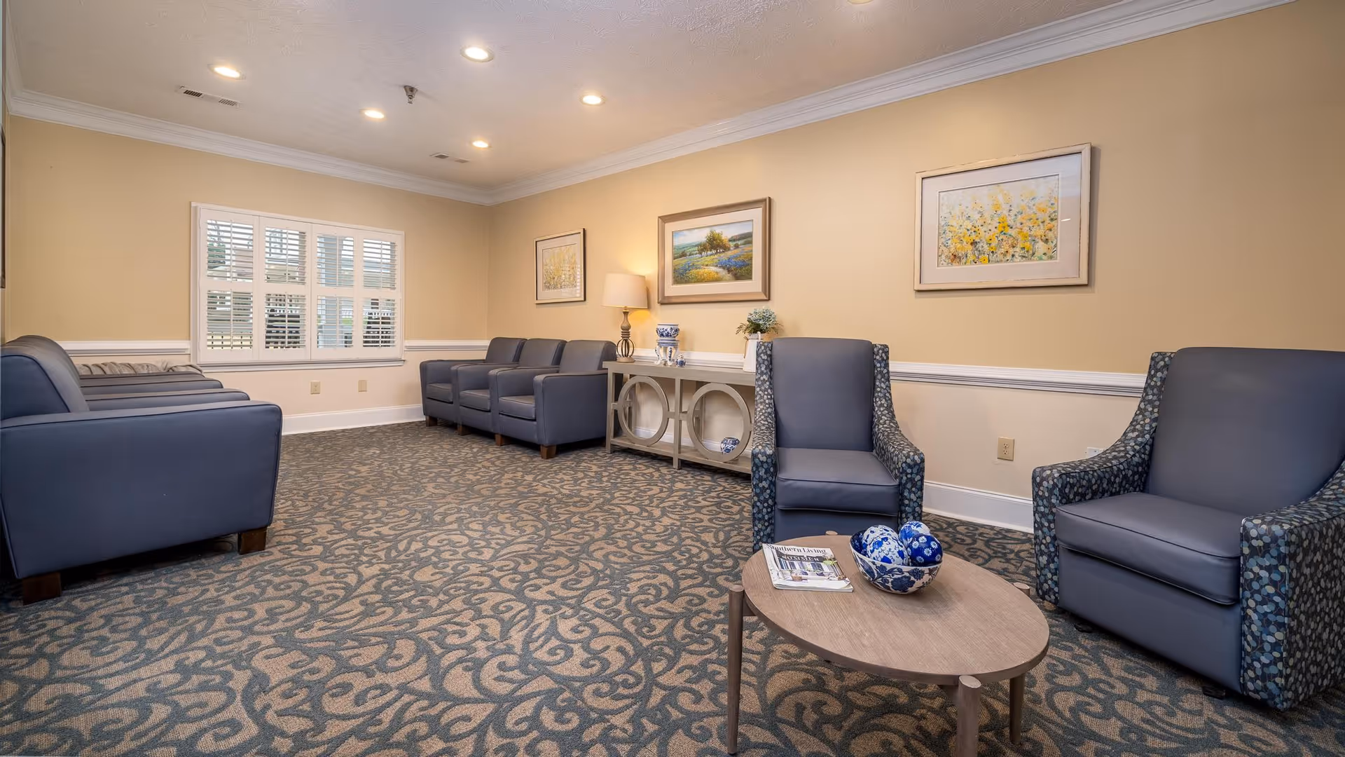 A well-lit living room area with patterned carpet, beige walls, and recessed ceiling lights. The room features multiple blue armchairs arranged along the walls and around a small wooden coffee table with decorative blue and white bowls and magazines. There is a console table against the wall with a lamp, decorative items, and framed artwork hanging above it. A window with white shutters is visible on the left wall.
