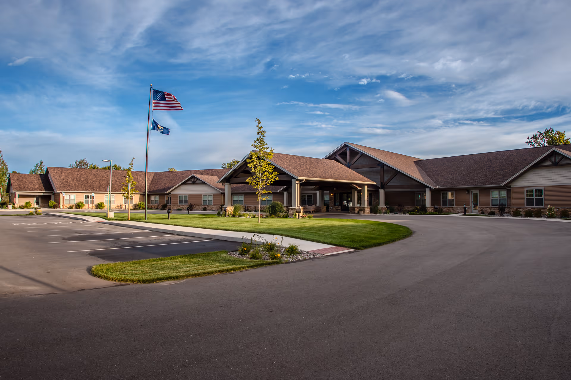 Exterior view of Teal Lake Senior Living Community building with a large covered entrance, two flagpoles with American and state flags, a parking lot, and well-maintained landscaping under a partly cloudy sky.
