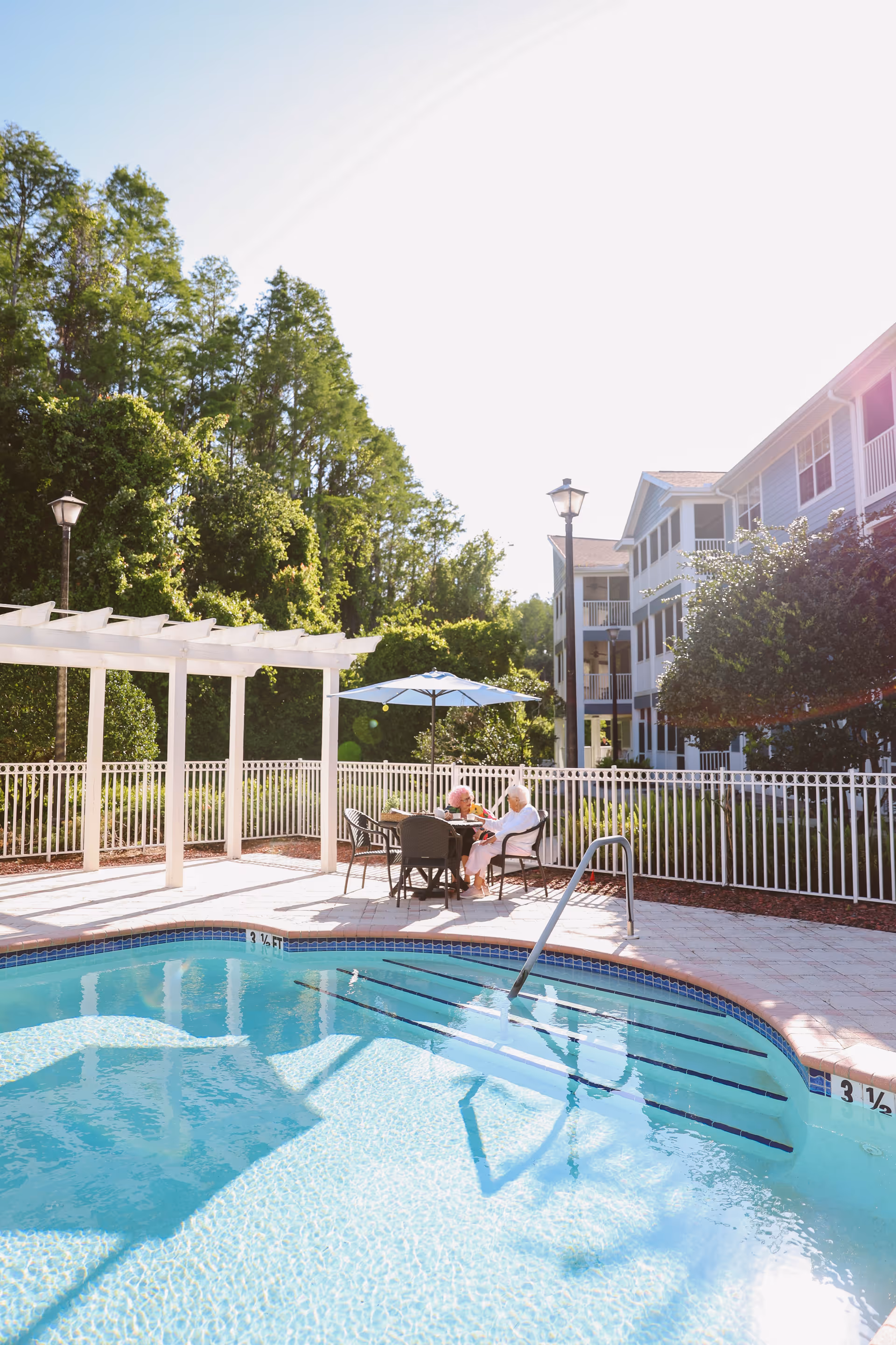 Outdoor pool area at a senior living facility with clear blue water, a white pergola, and a table with an umbrella where two elderly women are seated and conversing. The background shows a multi-story residential building and lush green trees.