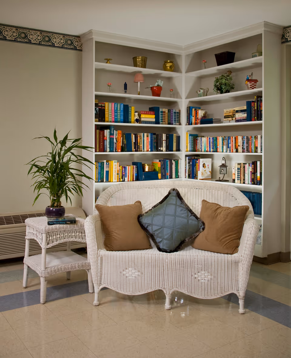 A cozy corner with a white wicker loveseat adorned with three cushions, positioned in front of a built-in bookshelf filled with various books and decorative items. To the left of the loveseat is a matching white wicker side table with a potted plant and a book on top. The floor is tiled and the walls are light-colored with a decorative border near the ceiling.