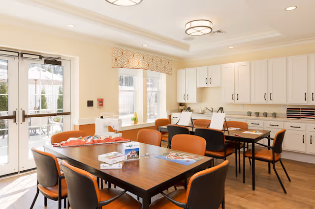 A bright and spacious activity room with large windows and glass doors letting in natural light. The room features two wooden tables surrounded by orange and black chairs. On the tables are art supplies, puzzles, and games. The back wall has white cabinets and countertops with small potted plants and organized trays. Outside the glass doors, there is a patio area with umbrellas and seating.
