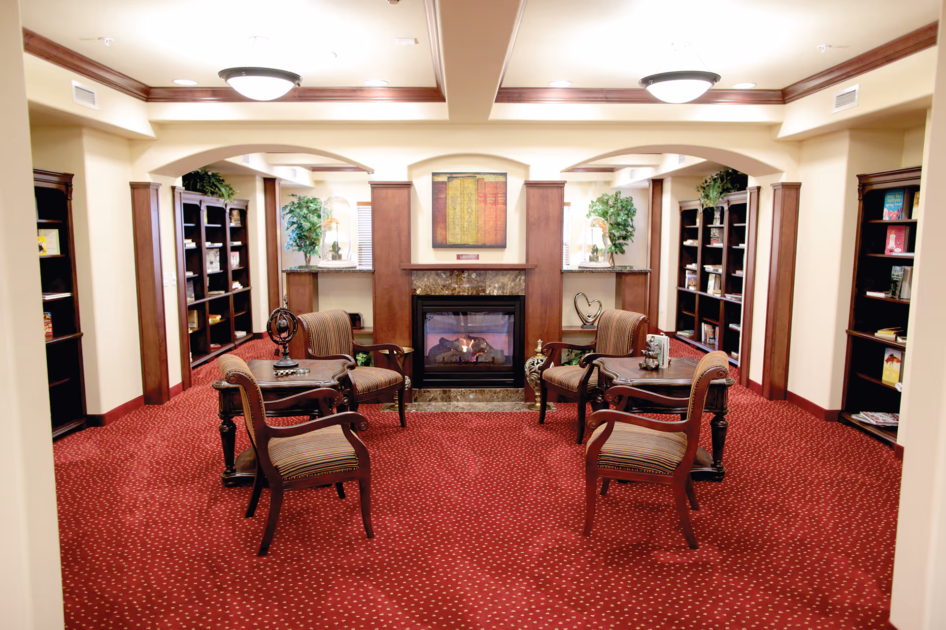 A cozy common area with a red carpet featuring small white dots, four wooden armchairs with striped cushions arranged around two small wooden tables, and a fireplace with a marble surround. The room has built-in dark wood bookshelves lining the walls, decorative plants, and warm lighting from ceiling fixtures.