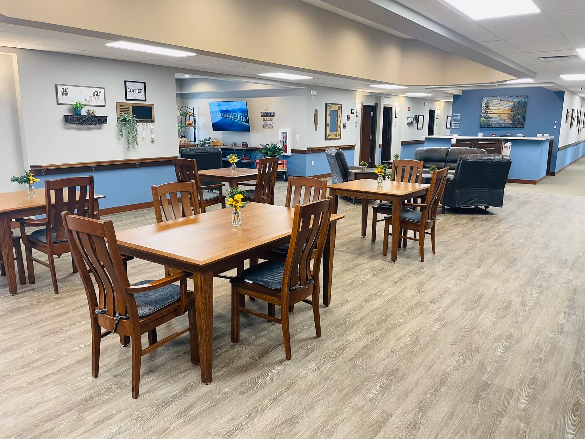 A spacious senior living common area with wooden tables and chairs arranged neatly. Each table has a small vase with yellow flowers. In the background, there are comfortable black leather chairs and a mounted flat-screen TV on the wall. The walls are painted in light gray and blue tones, decorated with framed pictures and small shelves with plants and decor items. The floor has a light wood finish.
