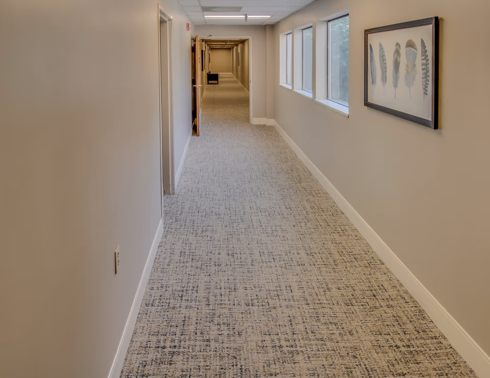 A long, carpeted hallway in a senior living facility with beige walls, several windows on the right side, and framed artwork featuring feathers hanging on the wall. The hallway extends into the distance with doors and a chair visible at the far end.