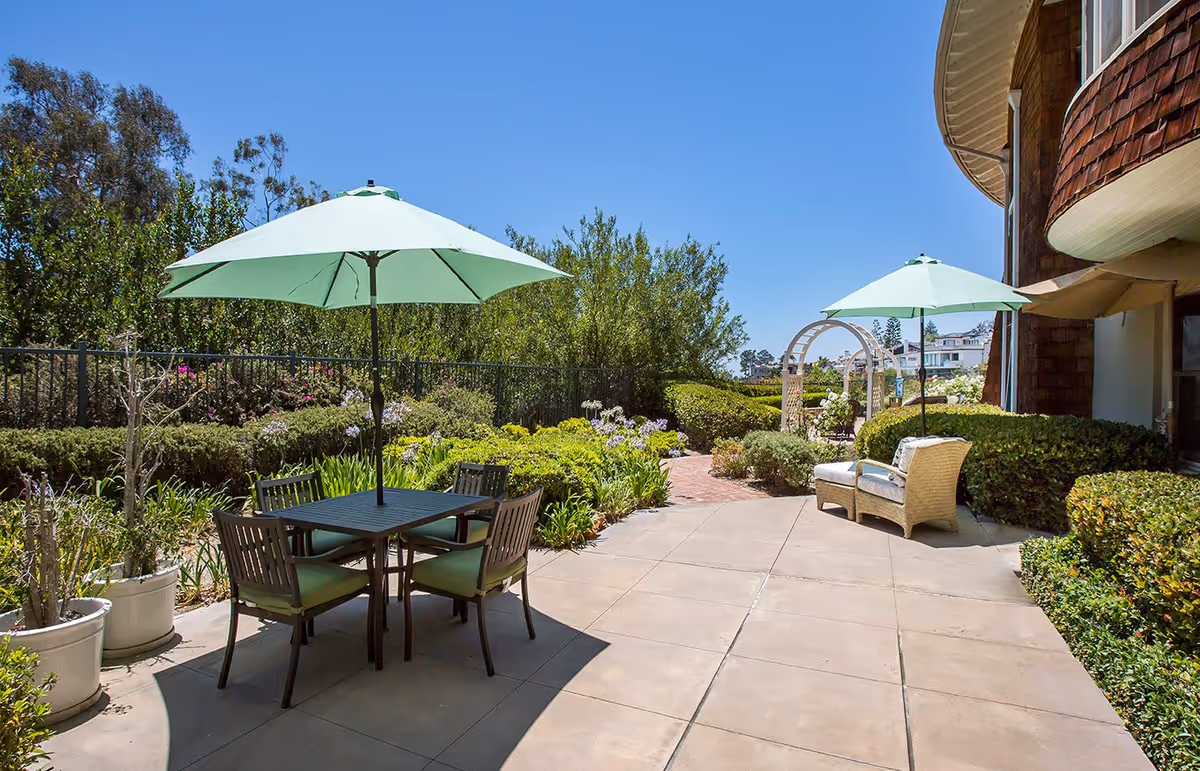 Outdoor patio area with two green umbrellas providing shade over a table with four chairs and a pair of cushioned wicker chairs. The patio is surrounded by lush greenery, flowering plants, and a white garden archway in the background under a clear blue sky.